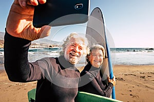 Couple of pensioners seniors taking a selfie together at the beach with their wetsuits and surfboards - mature people learning