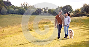 Couple With Pet Golden Retriever Dog Walking Along Path Across Field In Countryside