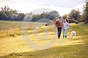 Couple With Pet Golden Retriever Dog Walking Along Path Across Field In Countryside