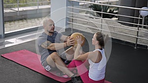 The couple pf sportsmen synchronically does the sit-ups and hands over the ball in the gym.