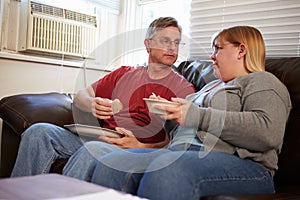 Couple With Poor Diet Sitting On Sofa Eating Meal