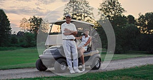 Couple posing golf cart outside. Two golfers take clubs sport equipment on field