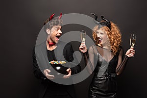 Couple posing in halloween costumes with glasses of champagne and pot of candies on black