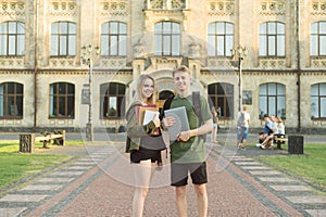 Couple of positive happy students standing at college entrance holding notebooks, books and laptop. Two cheerful attractive young