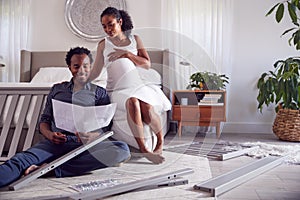 Couple With Pregnant Woman Looking At Instructions For Self Assembly Baby Cot