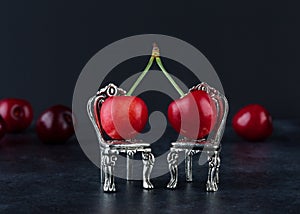 Couple of red cherries on two silver chairs with blurred cherries in the black backround