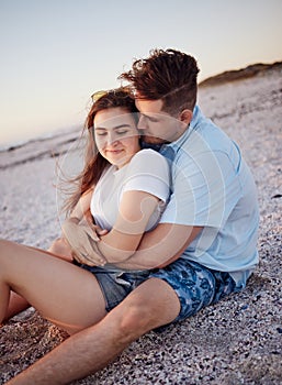 Couple, relax and hug for love on a beach sitting and enjoying time together in the outdoors. Young man hugging woman