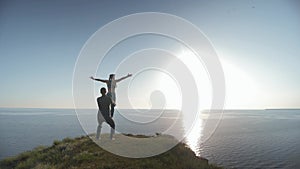 Couple relaxing by practicing yoga on mountain near calm sea with sunlight, athletic girl is balancing on legs of her