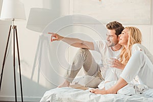 couple relxing in bed with cup of coffee and pointing at window