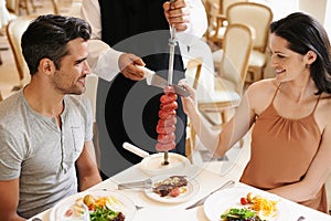 Couple, restaurant and waiter serving food or sausage kebab on skewer for fine dining, anniversary or nutrition. Man