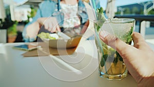 A couple is resting in a cafe. First person view - a man holding a glass with a cocktail, a woman in the background