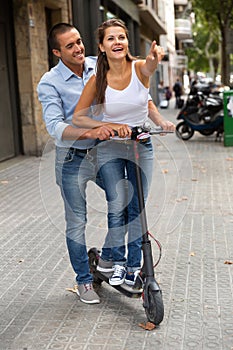Couple riding kick scooter together, woman pointing to sightseeing