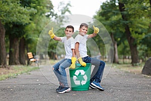 A couple of schoolers in yellow latex gloves sitting on a recycling bin and showing thumbs up on a blurred park