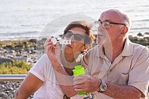 Couple of seniors married sit on the beach blowing a bubble of soap with the sea in the background - having fun together