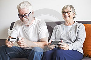 Couple of seniors sitting on the sofa and playing video games holding a controller - people have fun and enjoy  together laughing