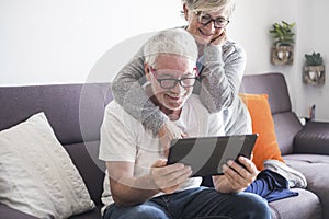 Couple of seniors smiling and looking at the same tablet hugged on the sofa - indoor, at home concept - caucasians mature and