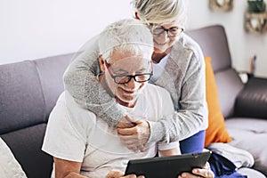 Couple of seniors smiling and looking at the tablet - woman hogging at man with love on the sofa - indoor