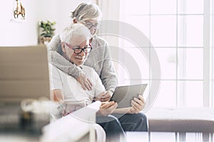 Couple of seniors smiling and looking at the tablet - woman hogging at man with love on the sofa - indoor