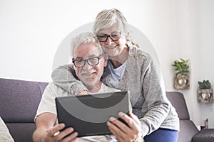 Couple of seniors smiling and looking at the tablet - woman hogging at man with love on the sofa - indoor - showing