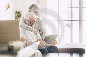 Couple of seniors smiling and looking at the tablet - woman hogging at man with love on the sofa - indoor - showing