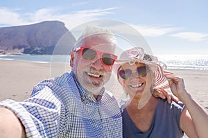 Couple of seniors travelling and taking a selfie in a beautiful beach with a mountain at the background - smiling and ejoying