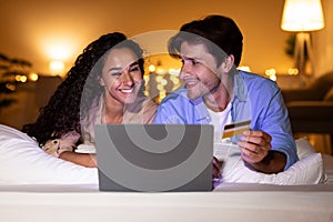 Couple Shopping Online Using Laptop And Credit Card In Bedroom