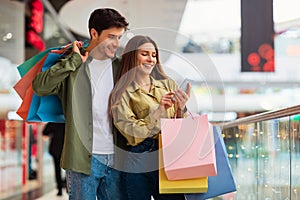 Couple Shopping Using Cellphone Holding Shopper Bags Standing In Mall