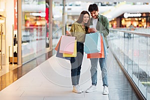 Couple Shopping Using Smartphone Application Holding Shopper Bags In Hypermarket