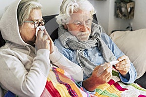 Couple of sicks seniors and mature people sitting at the sofa with fever looking at the thermometer with high temperature - two