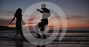 Couple, silhouette and playing on beach with water splash for fun outdoor evening or bonding together. Man and woman