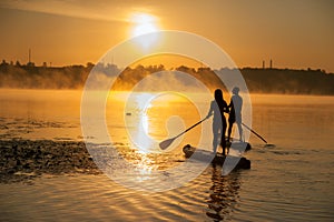 Couple silhouette on surfboards on the water at sunrise, fog on the horizon. SUP surfing at sunset