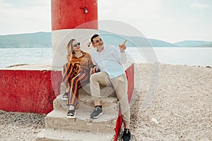 A couple sits near a lighthouse on the beach against the backdrop of the sea and mountains. Vacation love, honeymoon, travel.