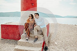 A couple sits near a lighthouse on the beach against the backdrop of the sea and mountains. Vacation love, honeymoon, travel.