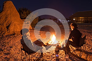 Couple sitting at burning camp fire in the night. Camping in the desert with wild elephants in background. Summer adventures and e