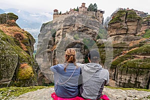 Meteora - Couple sitting on cliff edge with scenic view of Holy Monastery of Great Meteoron appearing from fog, Kalambaka, Meteora