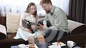 Couple is sitting on the couch sofa at home Look into the tablet and smile, Drink tea from a white tea set and watching