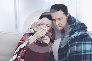 The couple is sitting on the couch wrapped in blankets. Man and woman are sick. A man hugs a woman who flies
