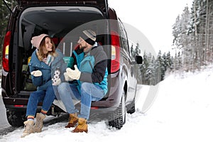 Couple sitting in open car trunk full of luggage near road, space for text. Winter