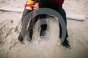 Couple sitting on the sand at winter beach. Half-length portrait. Couple's legs in hipster shoes close up