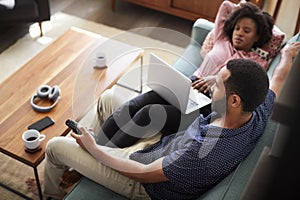 Couple Sitting On Sofa At Home Using Laptop Computer And Watching TV