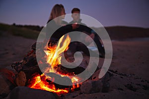 Couple Sitting On Surfboard By Camp Fire On Beach Using Mobile Phone As Sun Sets Behind Them