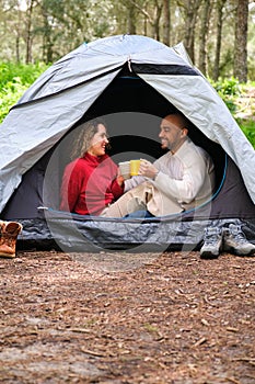 A couple is sitting in a tent, holding cups of coffee and smiling.