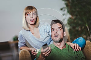 Couple sitting together on a sofa at home watching television, joyfully smiling enjoying a night in together