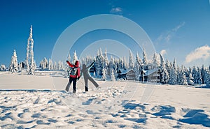 Couple in ski suits with backpack looking at beautiful view with snowy fir trees covered by snow in frozen mountains landscape.