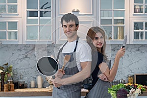 Couple standing back to back in kitchen. Smiling handsome young man wear an apron while holding frying pan and wooden spoon