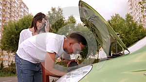 A couple is standing near a broken car with an open hood in the parking lot.