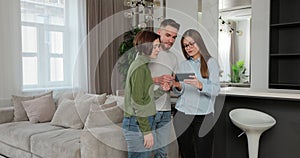 Couple standing in new house talking to woman real estate sales manager who is using tablet showing details. Buying home