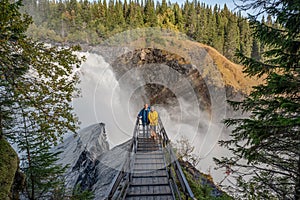 Couple standing on viewpoint bridge waterfall Tannforsen northern Sweden, rainbow in mist rapid flowing cascades water