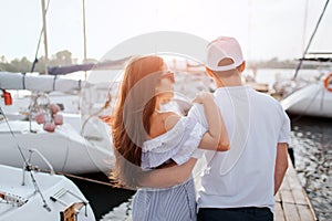 Couple stands and look on yachts that are on each sode from pier. They hug each other. Girl wears dress and sunglasses