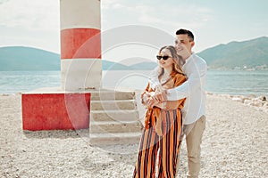 A couple stands near a lighthouse on the beach against the backdrop of the sea and mountains. Vacation love, honeymoon, travel.